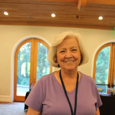 Smiling elderly woman with white hair wearing a purple shirt and conference badge standing indoors.