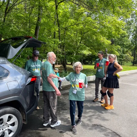 Group of six people casually gathered near a car on a sunny day, wearing green shirts and holding red cups outdoors.
