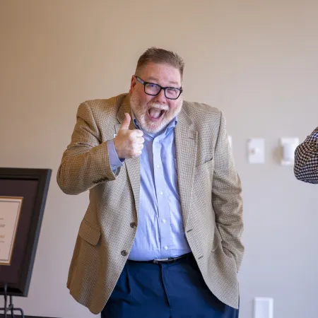Man in beige blazer giving thumbs up and smiling next to a man in checkered shirt with arms crossed indoors