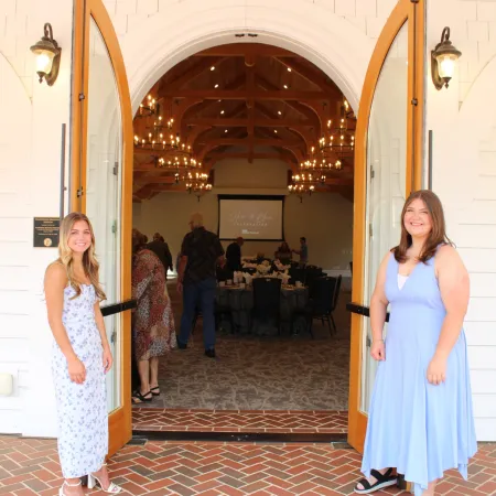 Two women in dresses smiling at the camera standing outside a warmly lit venue with chandeliers and wooden beams.