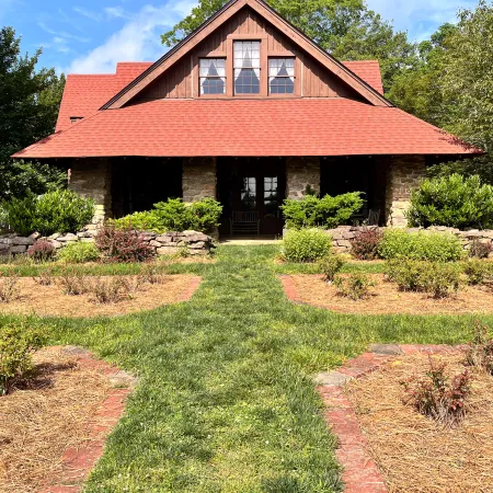 Rustic stone house with red roof surrounded by green lawn and landscaped garden beds under blue sky.