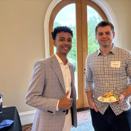 Two young men smiling indoors near arched wooden doors, one giving thumbs up, the other holding a plate of food.