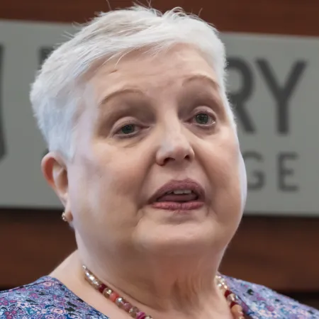 Older woman with short white hair wearing a beaded necklace and floral top in an indoor setting.