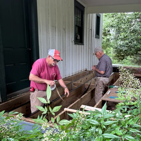 Two men working on a wooden deck attached to a white house surrounded by green plants and flowers.