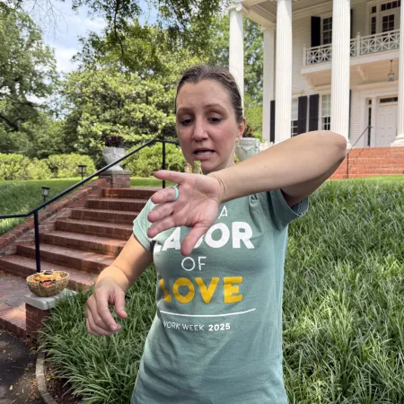 Woman wiping her face near brick steps and large white-columned house under green trees on a sunny day