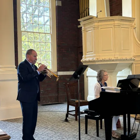 Man playing trumpet while woman accompanies on grand piano in a sunlit room with arched window.