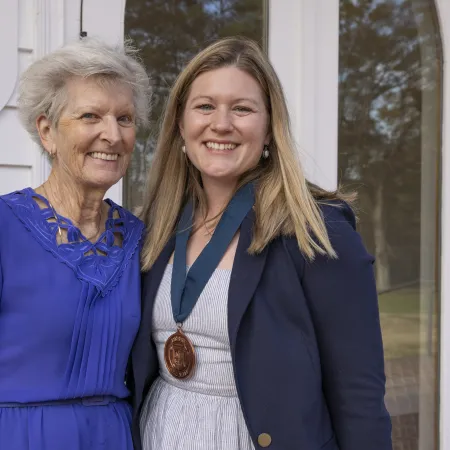Smiling elderly woman and young woman with medal standing together outside a white house entrance.