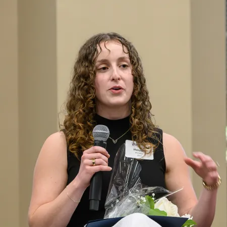 Young woman with curly hair speaking into a microphone while holding flowers and a folder at an event.