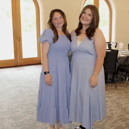 Two women in matching light blue dresses standing indoors near arched windows and wooden details in a bright room.