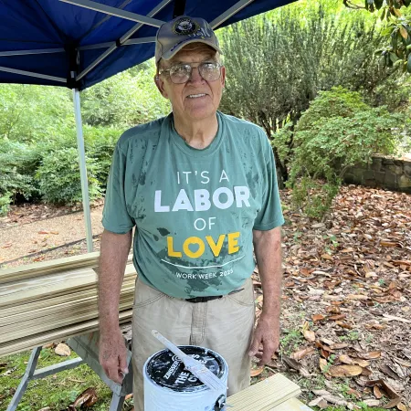 Elderly man in glasses and cap wearing a Labor of Love t-shirt stands outdoors under a blue canopy, holding paint supplies.