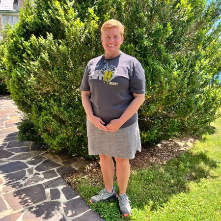 Smiling woman in gray t-shirt and skirt stands on stone path next to large green bush and lawn in sunny outdoor setting.