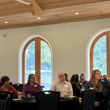 Group of adults attentively listening at a conference in a room with large arched windows and wooden ceiling beams.
