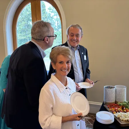Three people standing by a table with assorted vegetables and snacks, smiling and holding plates indoors.