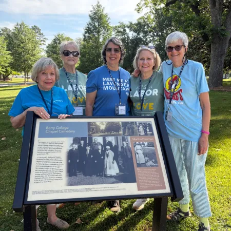 Group of five women smiling outdoors around a historical information sign on grass under trees in daylight