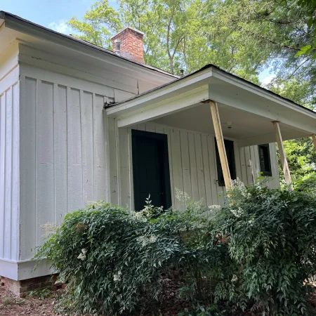 Small white wooden house with a green door, front porch supported by wooden beams, surrounded by green bushes and trees.