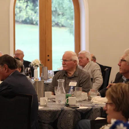 Senior adults seated at round tables during a daytime social or community event indoors near large windows.