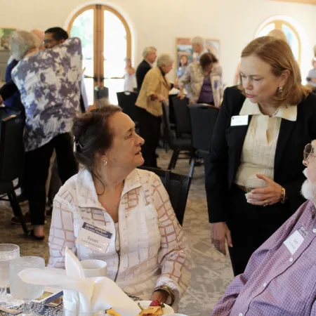 Three people engaged in conversation at a social event in a bright room with arched windows and seated guests.