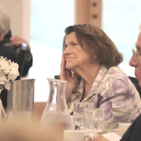 Elderly people sitting at a table with flowers, glasses, and a pitcher, engaged in a gathering or event.