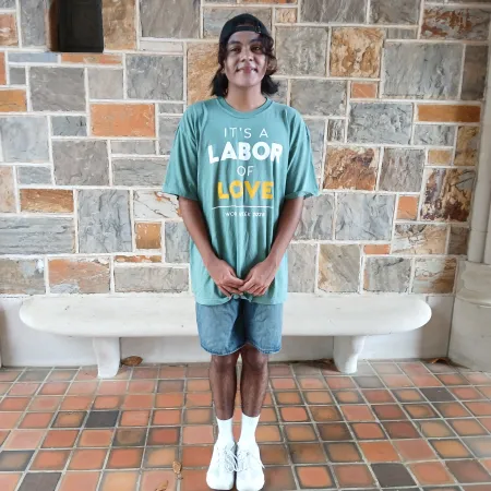 Teen boy wearing green shirt with labor of love message standing against stone wall in courtyard