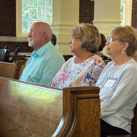 Four elderly people sitting in wooden pews inside a church with stained glass windows.