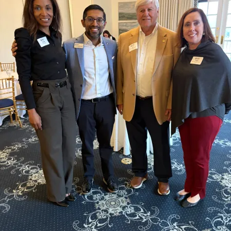 Four diverse professionals smiling indoors at a formal event on patterned carpet with name badges visible.