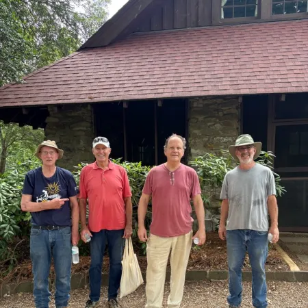 Four casually dressed men standing outdoors on a gravel path in front of a stone house with bushes and trees.