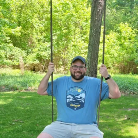 Man in blue t-shirt and shorts smiling on a swing in a sunny green park with trees in the background.