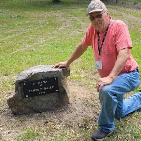 Senior man in cap kneeling beside memorial stone in a grassy park surrounded by trees