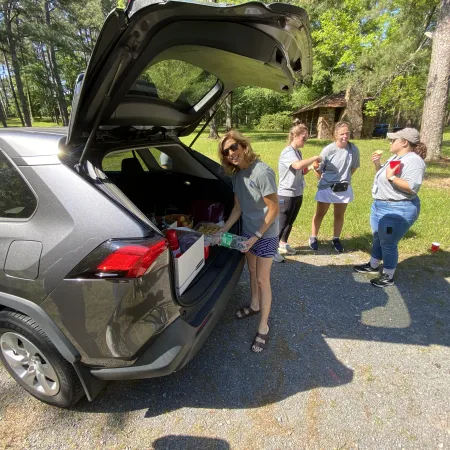 Group of women socializing outdoors near open SUV trunk on a sunny day with trees and grass in background