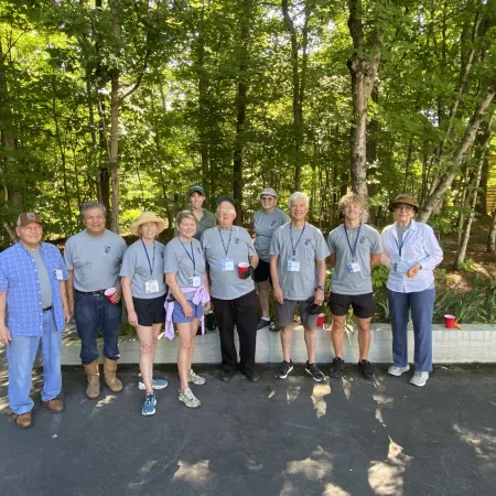 Group of people in casual attire standing outdoors in a wooded area on a sunny day