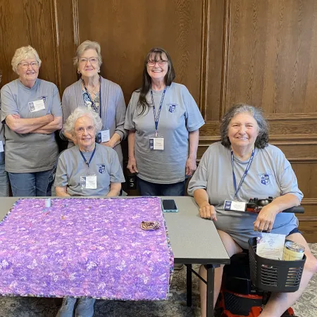 Group of six elderly women wearing matching gray shirts posing around a table with purple fabric on it.