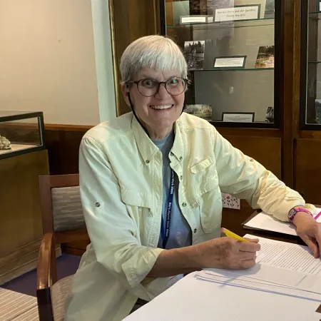 Smiling elderly woman with glasses writing on papers at a desk in a museum-like room with display cases.