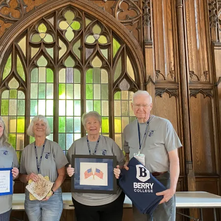 Four seniors wearing gray Berry College shirts holding crafts and framed art in a wooden chamber room with stained glass windows.