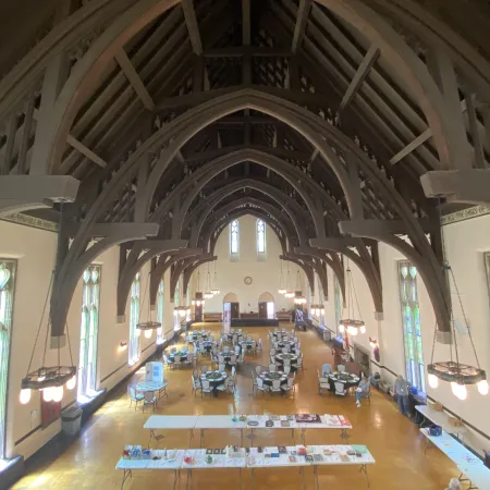 Spacious hall with wooden gothic arches, round tables, chairs, and hanging chandeliers arranged for event.