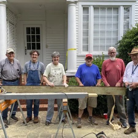 Group of six volunteers posing with a wooden plank and tools outside a white house with columns.