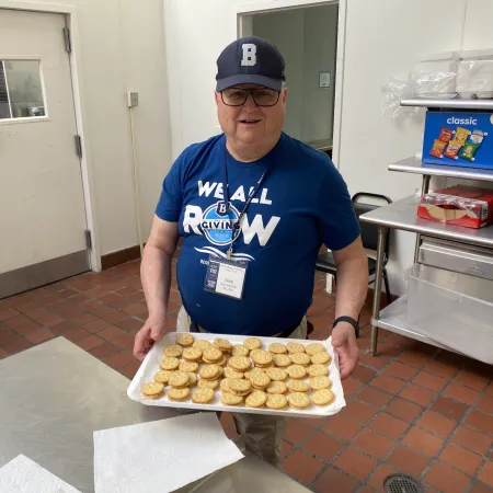 Man in blue shirt and cap holding tray of crackers in a kitchen with shelves and snacks.