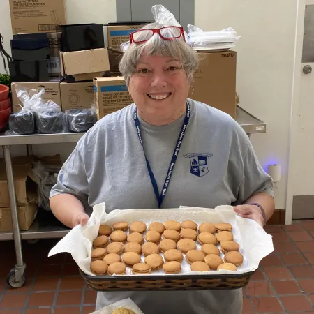 Smiling woman in gray shirt holding a tray of freshly baked cookies in a kitchen setting.