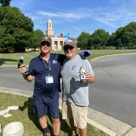 Two men smiling outdoors by a curved road near a building with a cupola, holding tools and spray paint cans.
