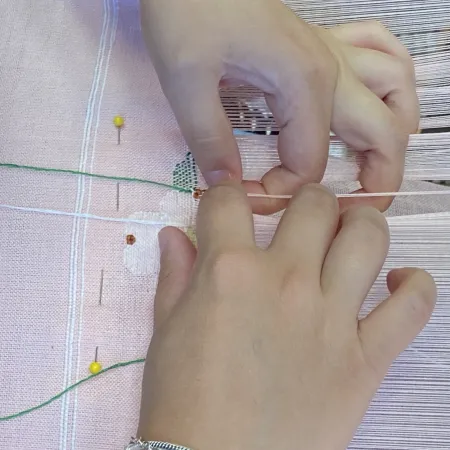 Hands weaving green thread on a pink fabric loom with pins and white threads creating a pattern.