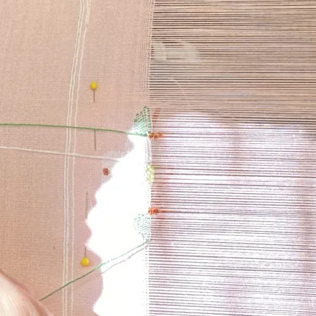 Close-up of a weaving loom with pink fabric and green thread secured by pins in natural light.