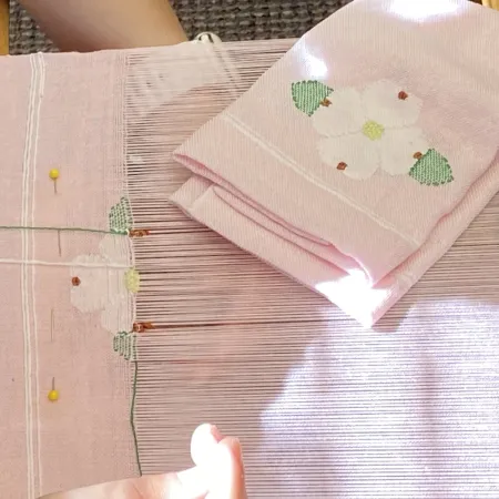 Pink fabric with embroidered white flowers in progress on a traditional weaving loom with pins and threads.