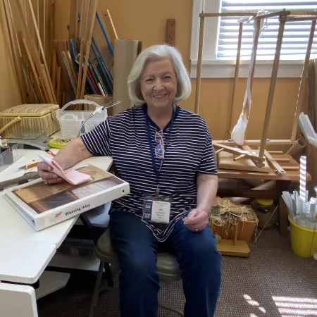 Smiling elderly woman sitting at a craft table with weaving supplies and tools in a sunny workshop.