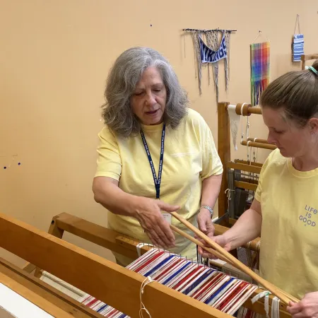 Two women in yellow shirts working collaboratively on a colorful weaving loom in a craft workshop.