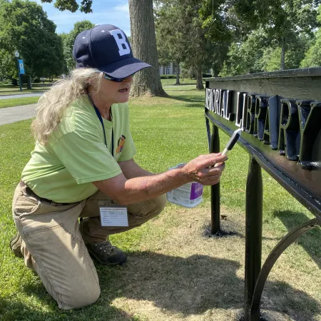 Person wearing a blue cap cleaning black metal library sign outdoors on a sunny day in a park.