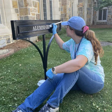 Young woman in blue cap painting black metal Alumni Center sign outside stone building on green lawn