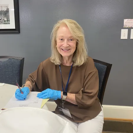 Smiling woman wearing blue gloves signs document at table in office with gray walls and framed picture.