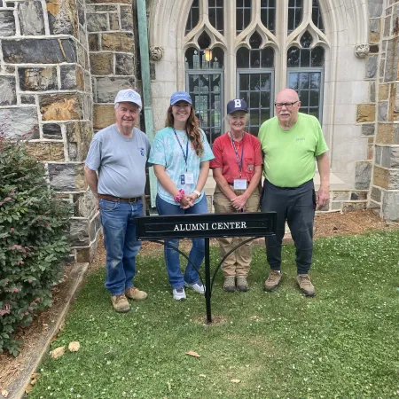 Four adults standing on grass outside a stone building with an Alumni Center sign in front of them.