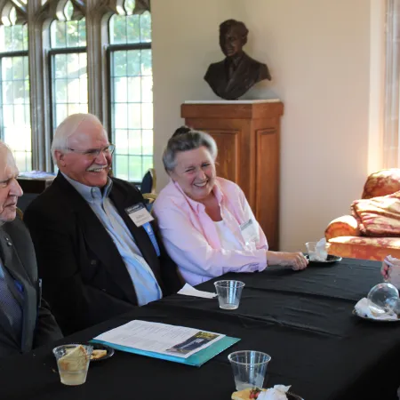 Three elderly people in formal attire sitting at a table with documents and drinks, engaged in a happy conversation.