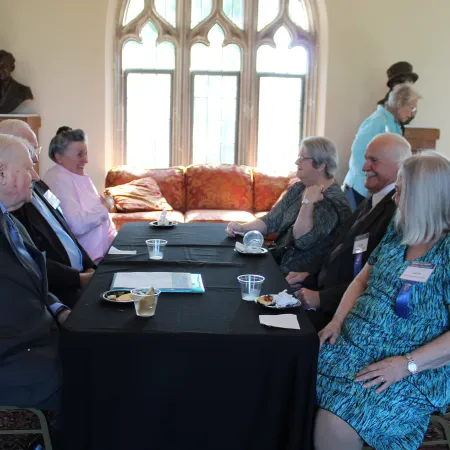 Group of elderly people sitting and chatting around a black table in a sunlit room with arched windows and bust statues.