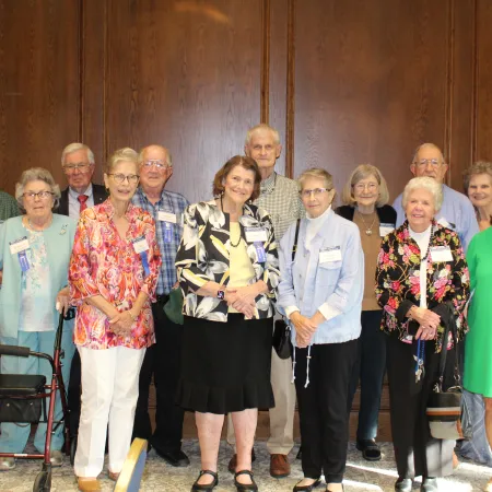 Group of elderly men and women standing indoors in front of wooden panel walls, smiling at the camera.